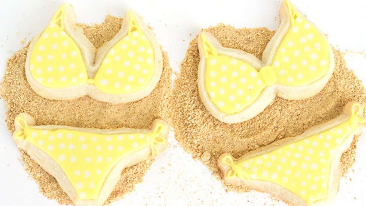 Four sugar cookies decorated as yellow polka dot bikinis rest on a layer of crushed cookie crumbs resembling sand—a playful treat perfect for spring desserts or fans of creative lemon desserts.