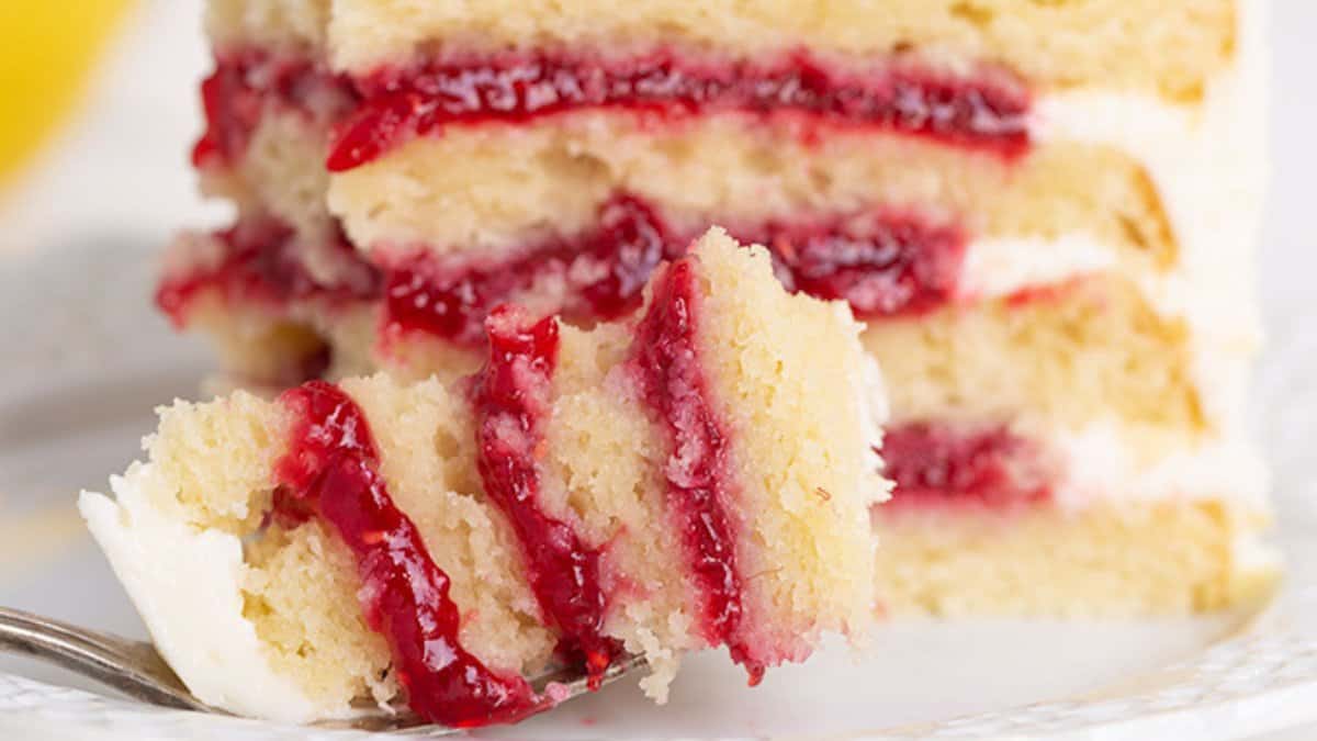 A close-up of a slice of layered cake with red fruit filling and white frosting, perfect for spring desserts, with a fork holding a bite-sized piece in the foreground.