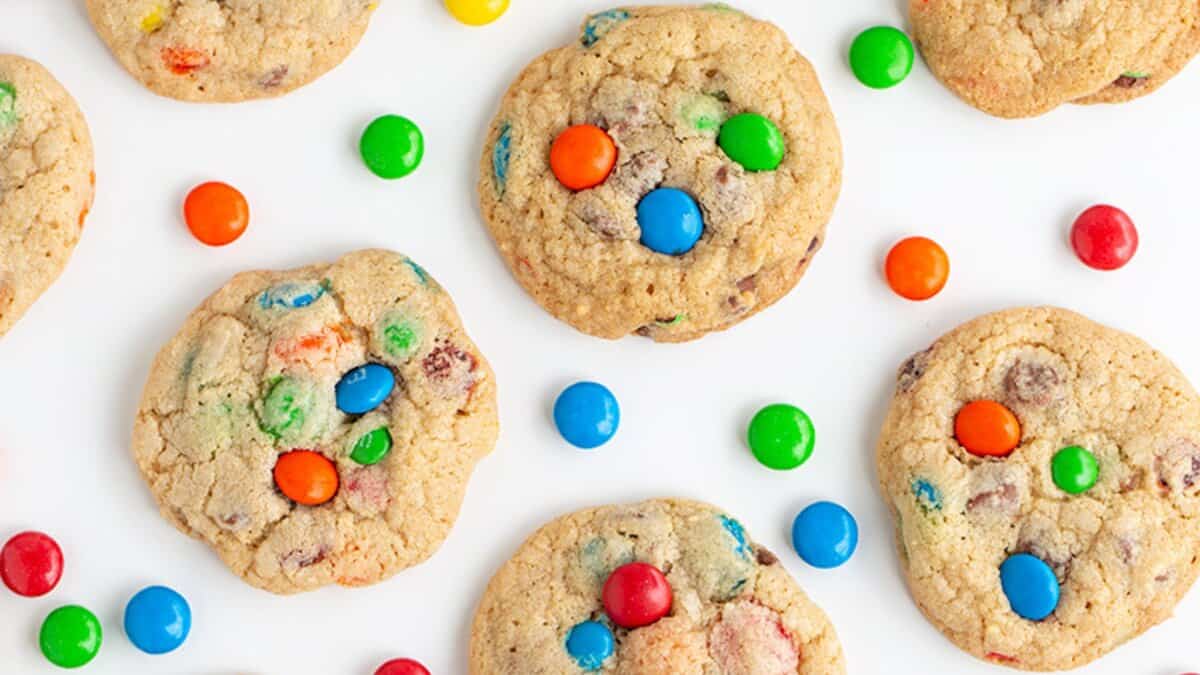 Cookies with colorful candy-coated chocolates are arranged on a white surface, perfect for trying out your favorite cookie recipes using pantry staples, with additional loose candies scattered around them.
