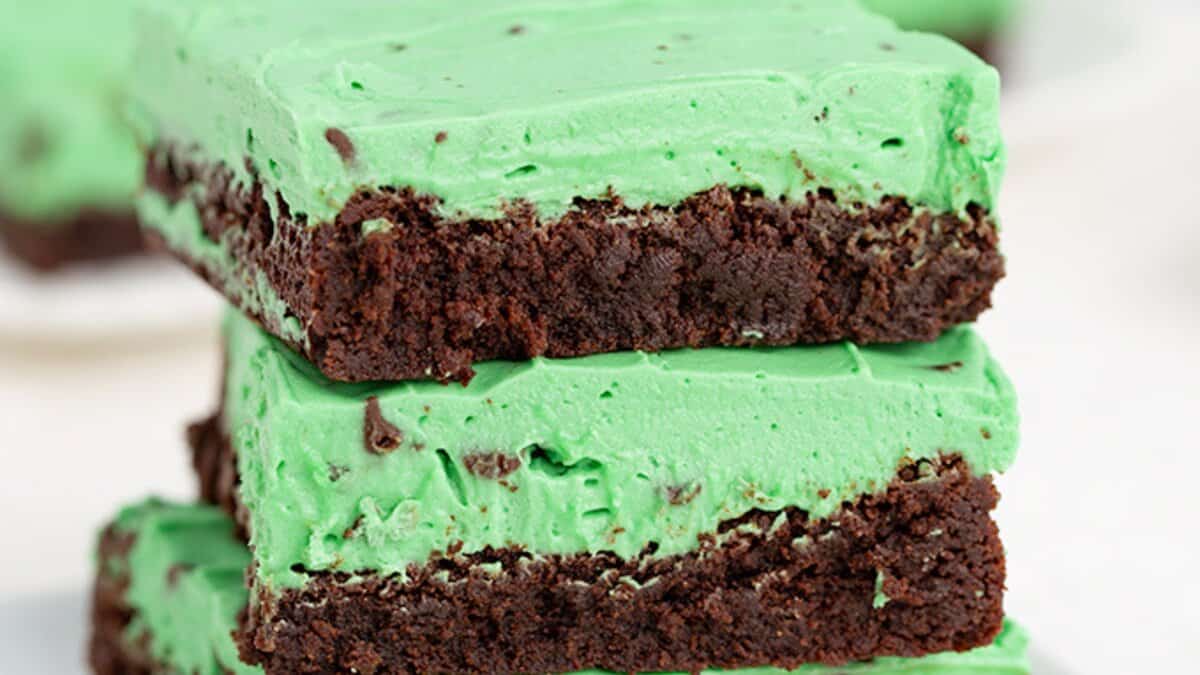 Three stacked chocolate brownies topped with a thick layer of green mint frosting and visible chocolate chips—an irresistible mint chocolate dessert, perfect for celebrating St. Patrick's Day.