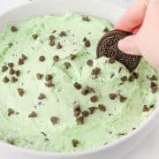 A hand dips a chocolate sandwich cookie into a bowl of Mint Chocolate Chip Dip, topped with chocolate chips.