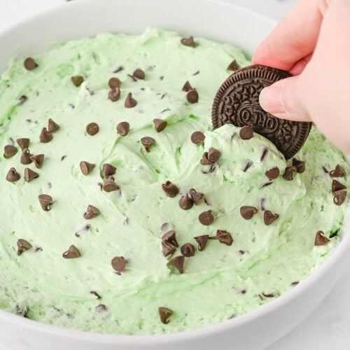 A hand dips a chocolate sandwich cookie into a bowl of Mint Chocolate Chip Dip, topped with chocolate chips.