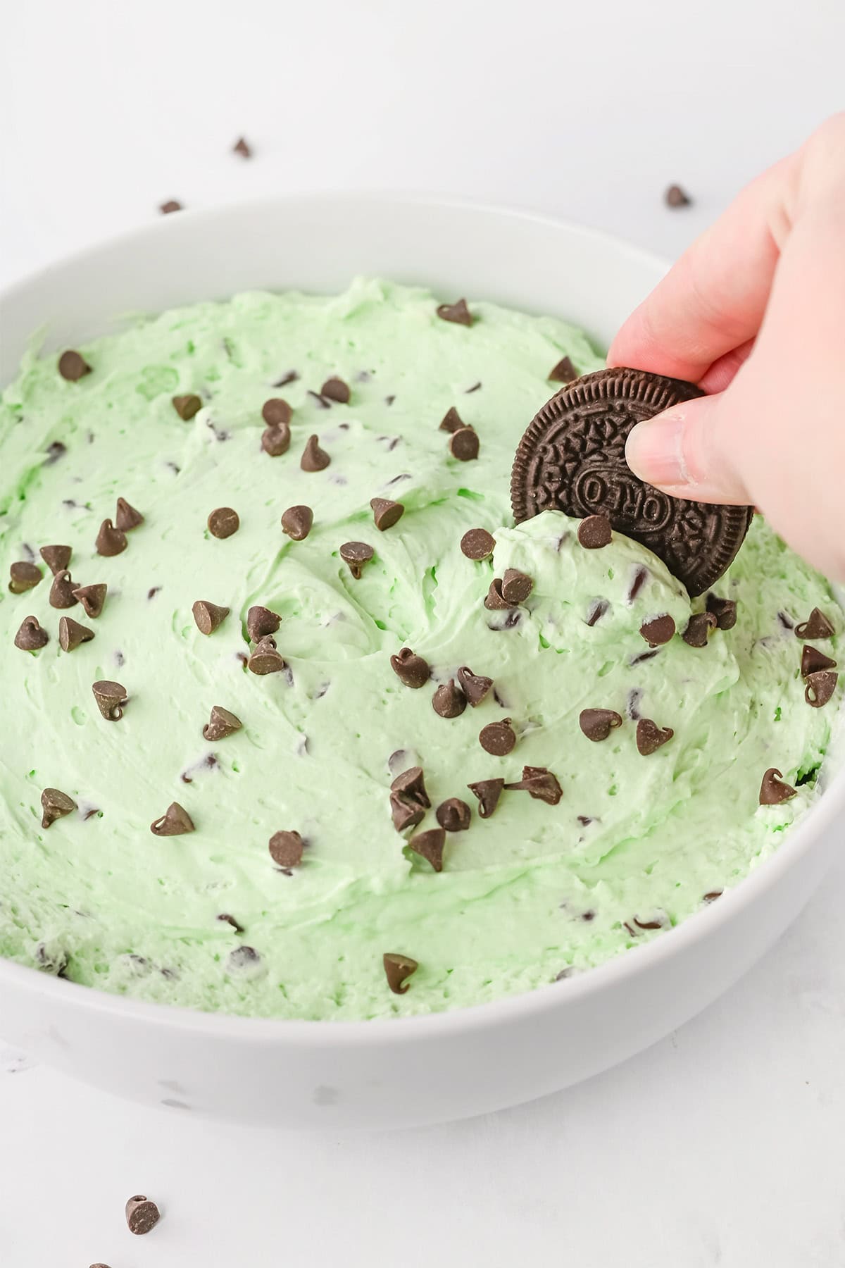 A hand dips a chocolate sandwich cookie into a bowl of Mint Chocolate Chip Dip, topped with chocolate chips.
