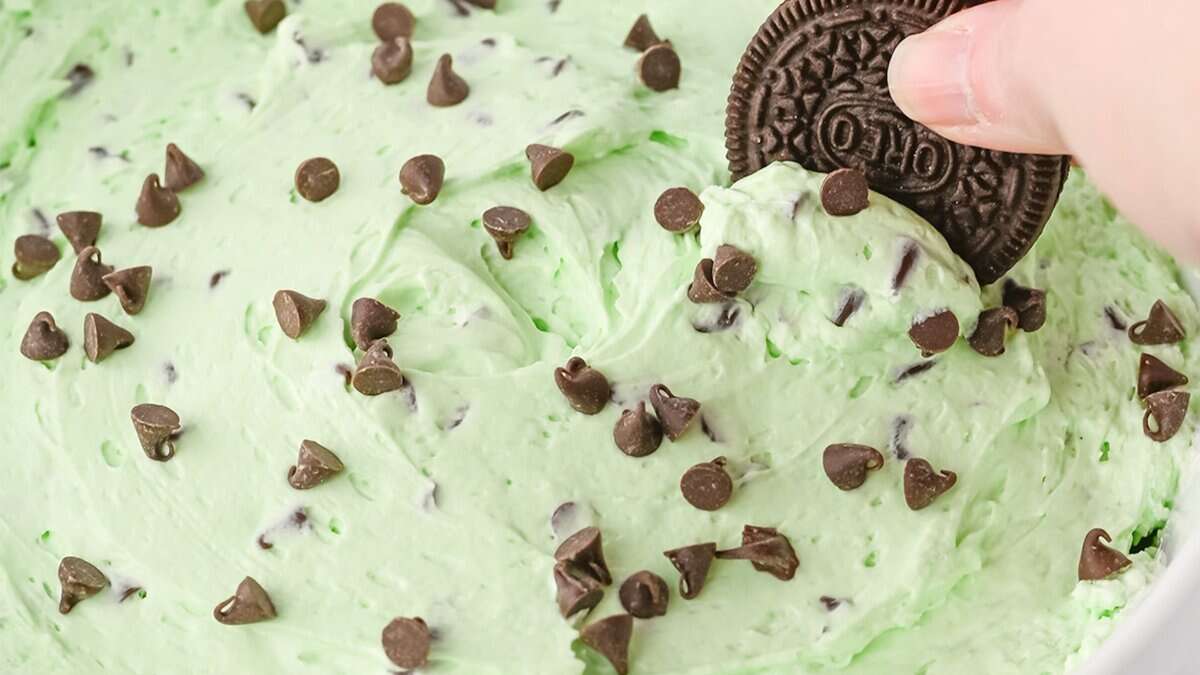 A hand dips a chocolate sandwich cookie into luscious mint chocolate chip dip, topped with chocolate chips—a fun twist on Mint Chocolate Desserts perfect for St. Patrick's Day.