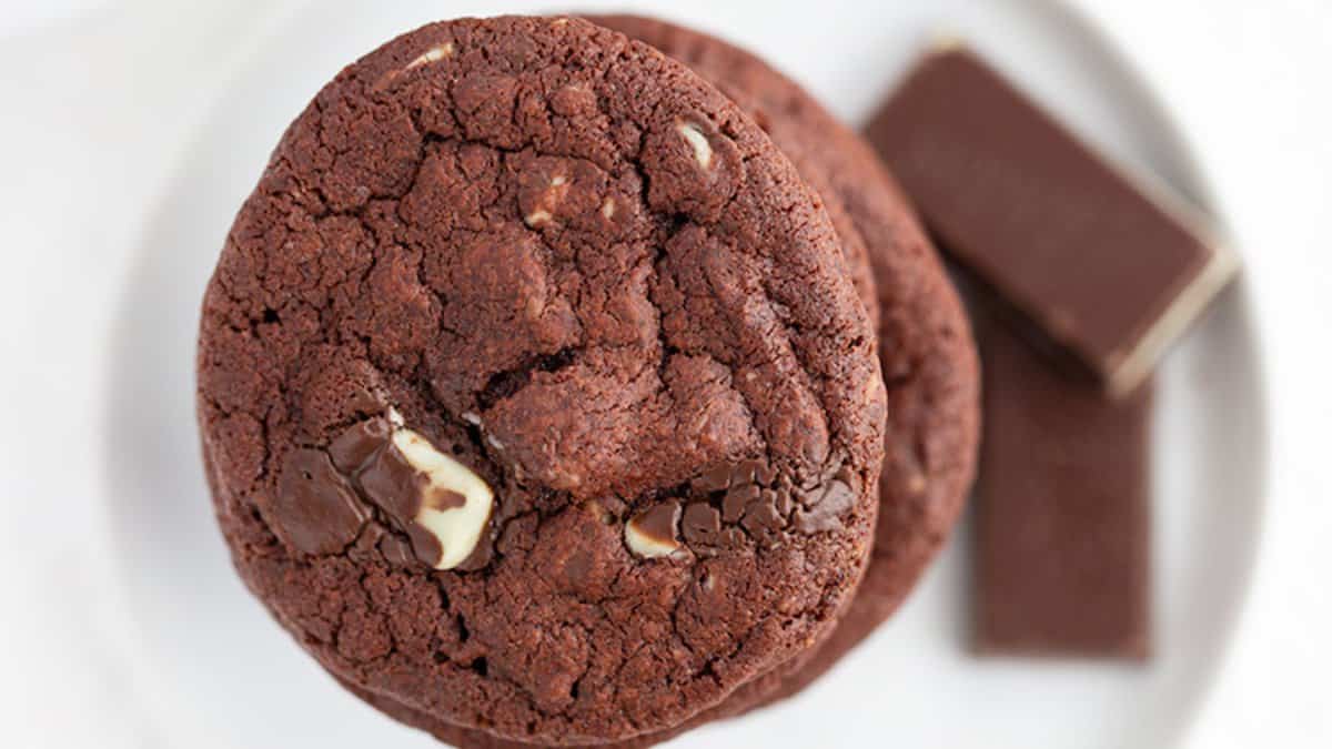 Two chocolate cookies with visible chocolate chunks are stacked on a white plate, with two pieces of chocolate next to them—a perfect treat for lovers of mint chocolate desserts.