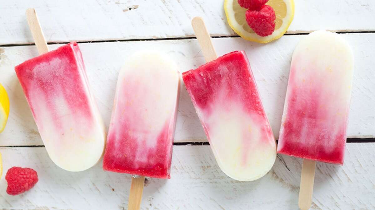 Four red and white popsicles on wooden sticks are arranged on a white wooden surface, with lemon slices and raspberries nearby—perfect for spring desserts or adding to your collection of delicious lemon recipes.