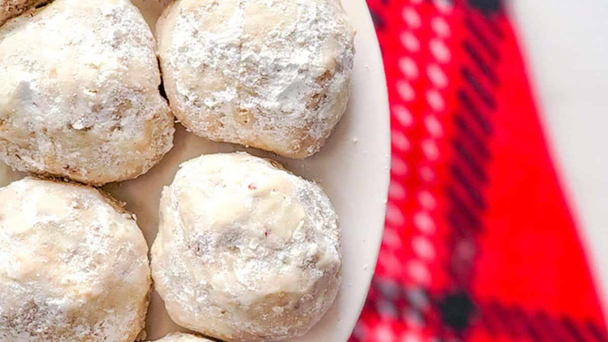 Six powdered sugar-coated cookies on a white plate, positioned next to a red and black patterned cloth&mdash;perfect for those seeking easy cookie recipes using pantry staples.