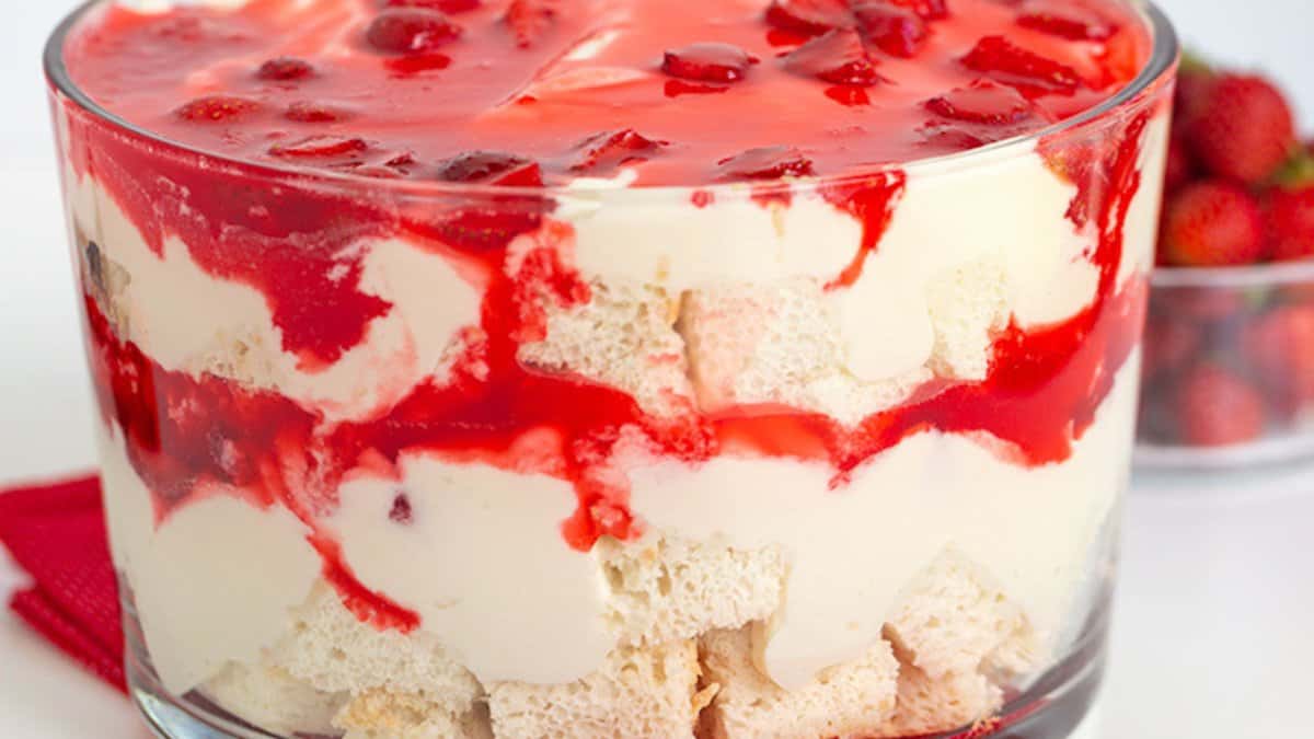 A glass bowl filled with layered strawberry dessert consisting of cake cubes, creamy filling, and strawberry topping—perfect for fans of fresh strawberry recipes. A small bowl of strawberries sits in the background.