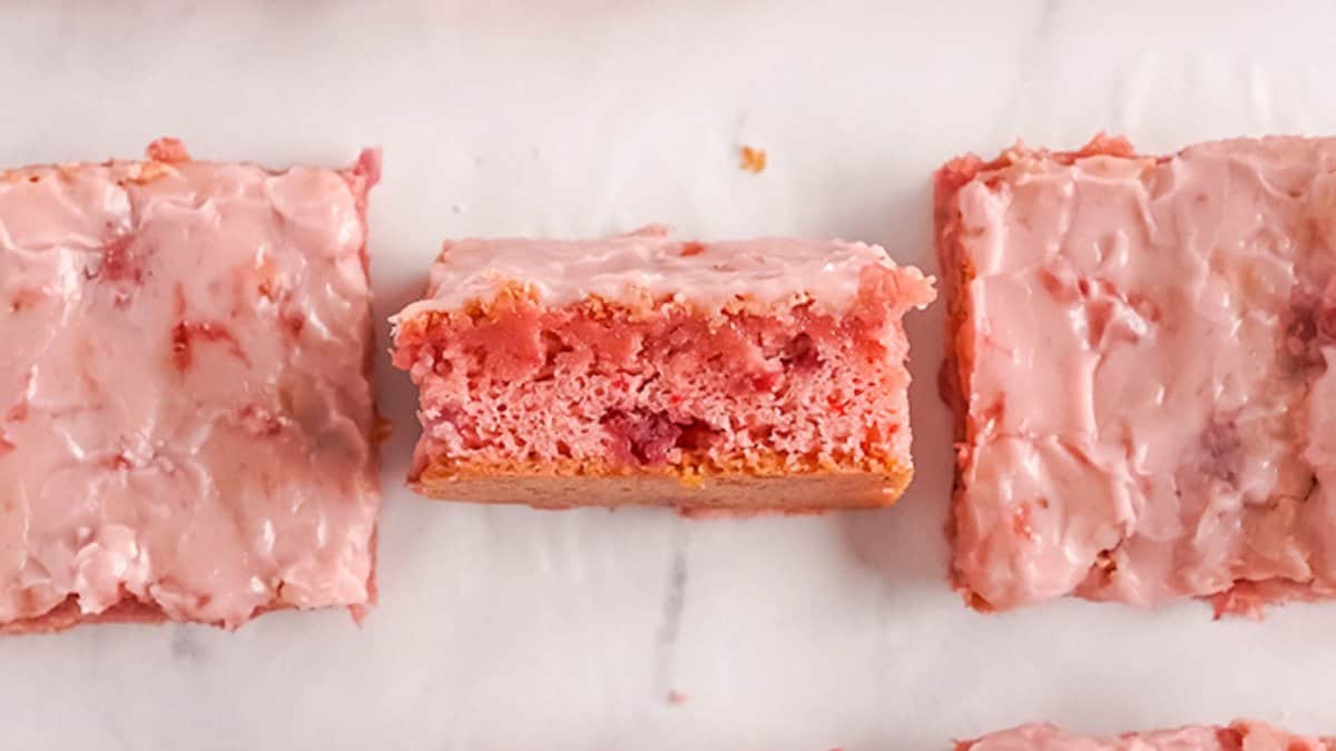 Rectangular strawberry cake bars with pink glaze are arranged on a white surface. One bar is cut to show the moist, pink interior with bits of fresh strawberry, making it a perfect spring dessert.