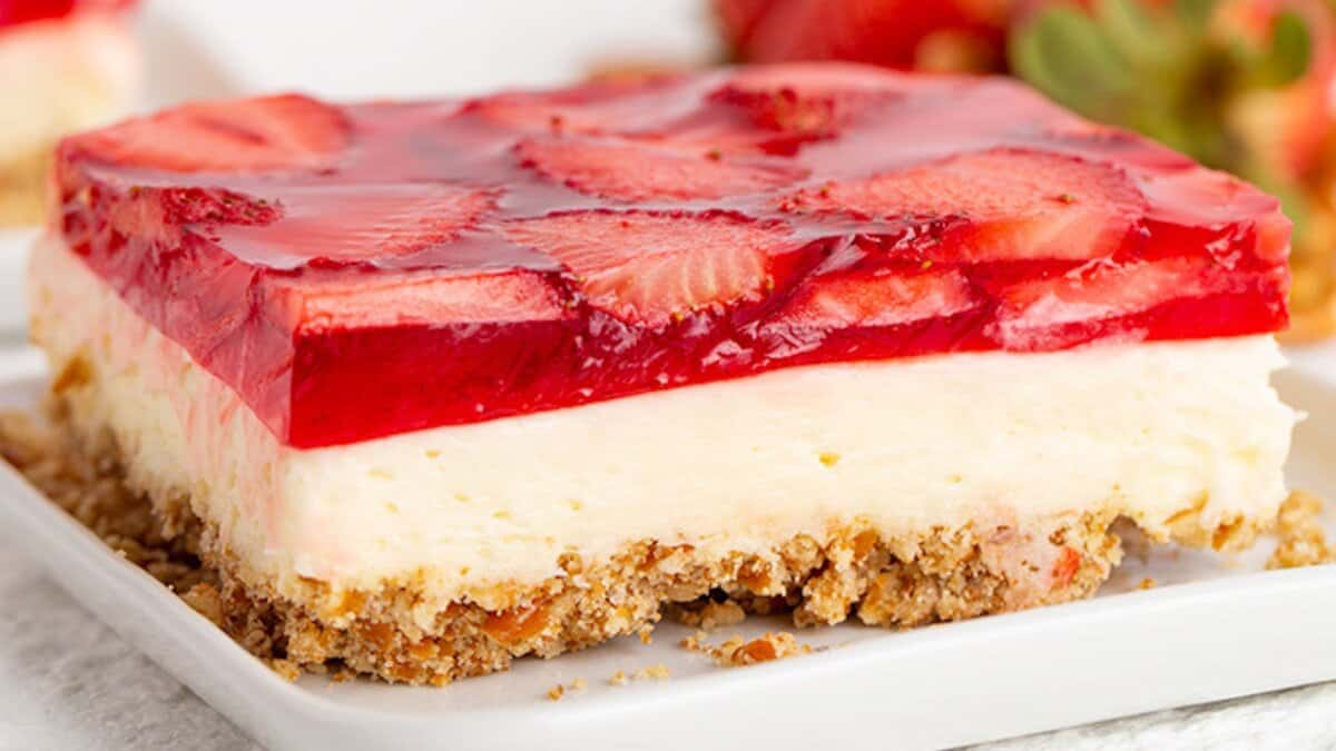 A slice of layered strawberry dessert with a crumbly crust, creamy middle layer, and fresh strawberry gelatin topping sits on a white plate—a perfect choice for spring desserts.