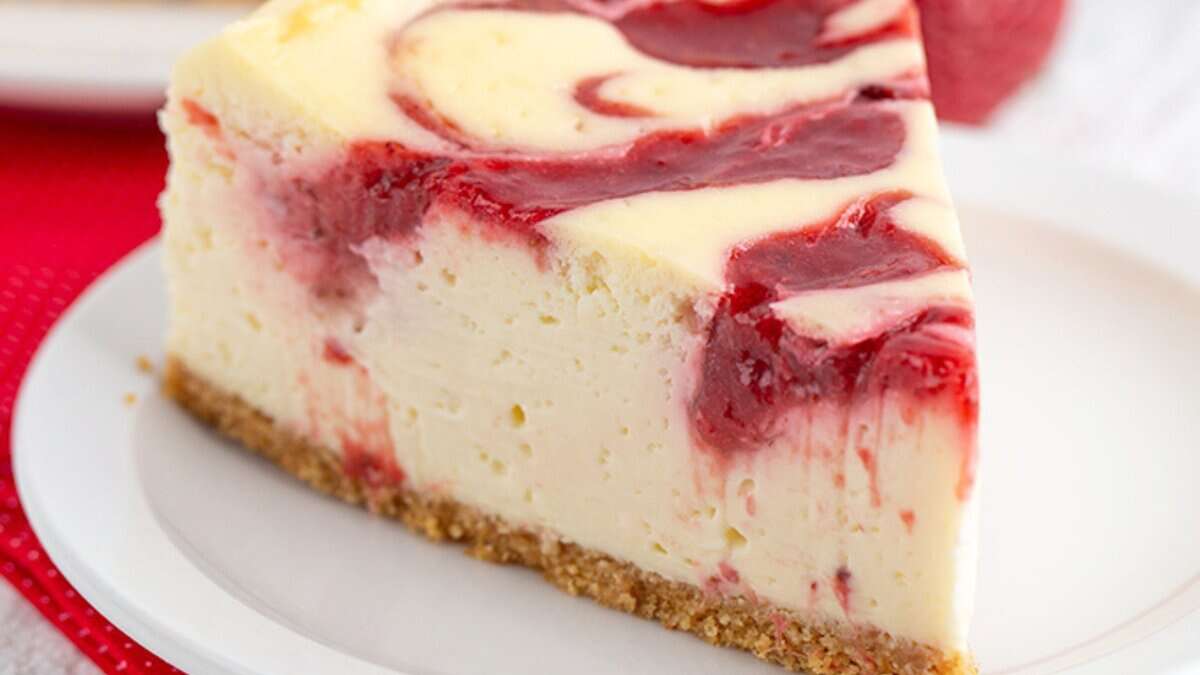 A slice of cheesecake with a graham cracker crust and raspberry swirl on a white plate, garnished with fresh strawberry for a perfect spring dessert.