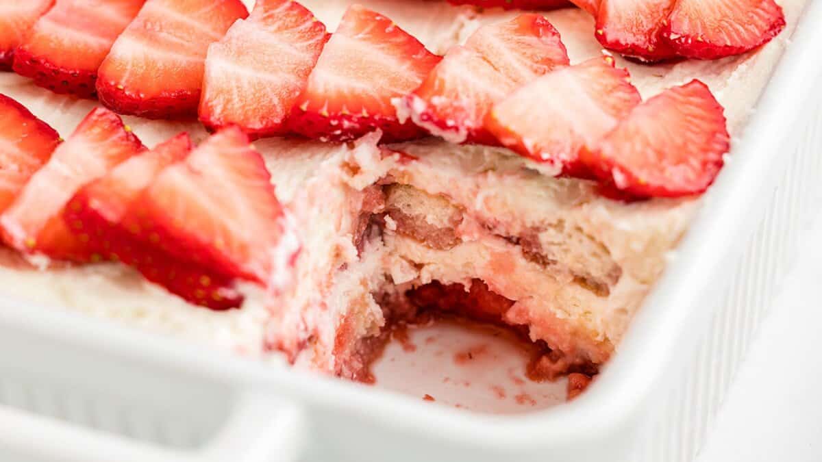 A close-up of a spring strawberry dessert in a white dish, topped with fresh strawberry slices, with a portion removed to reveal the layered cream and cake inside.