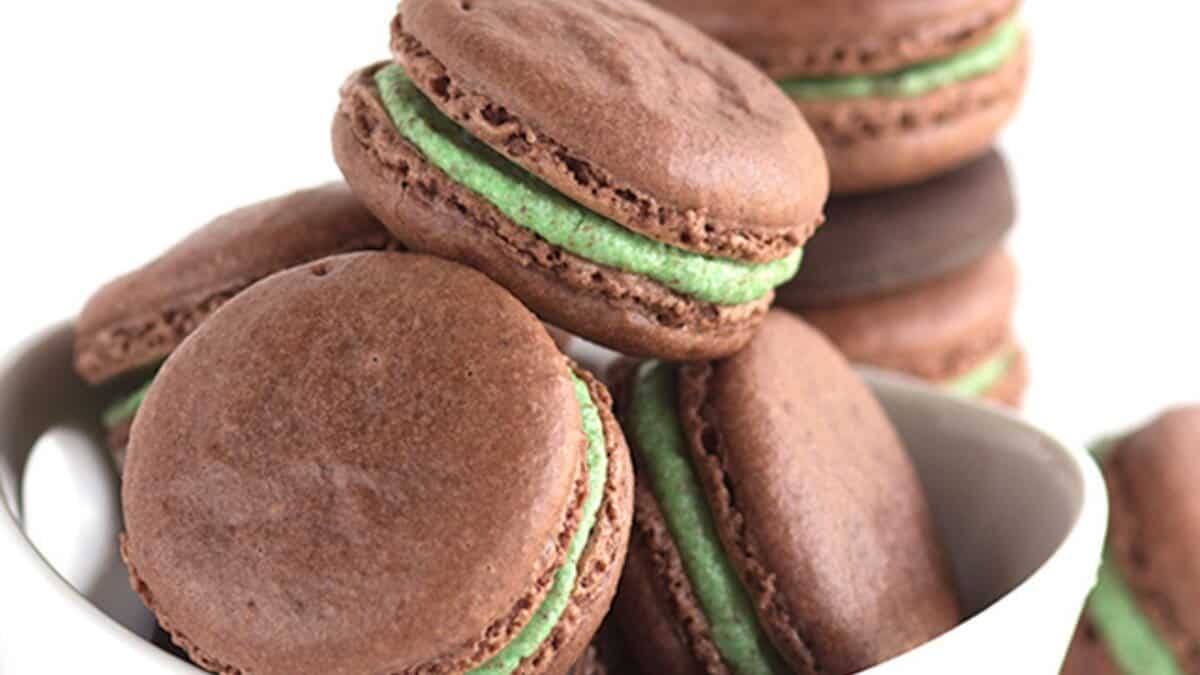 A bowl filled with chocolate macarons, each with a green mint chocolate filling, stacked on top of each other—perfect for celebrating St. Patrick's Day or enjoying mint chocolate desserts.