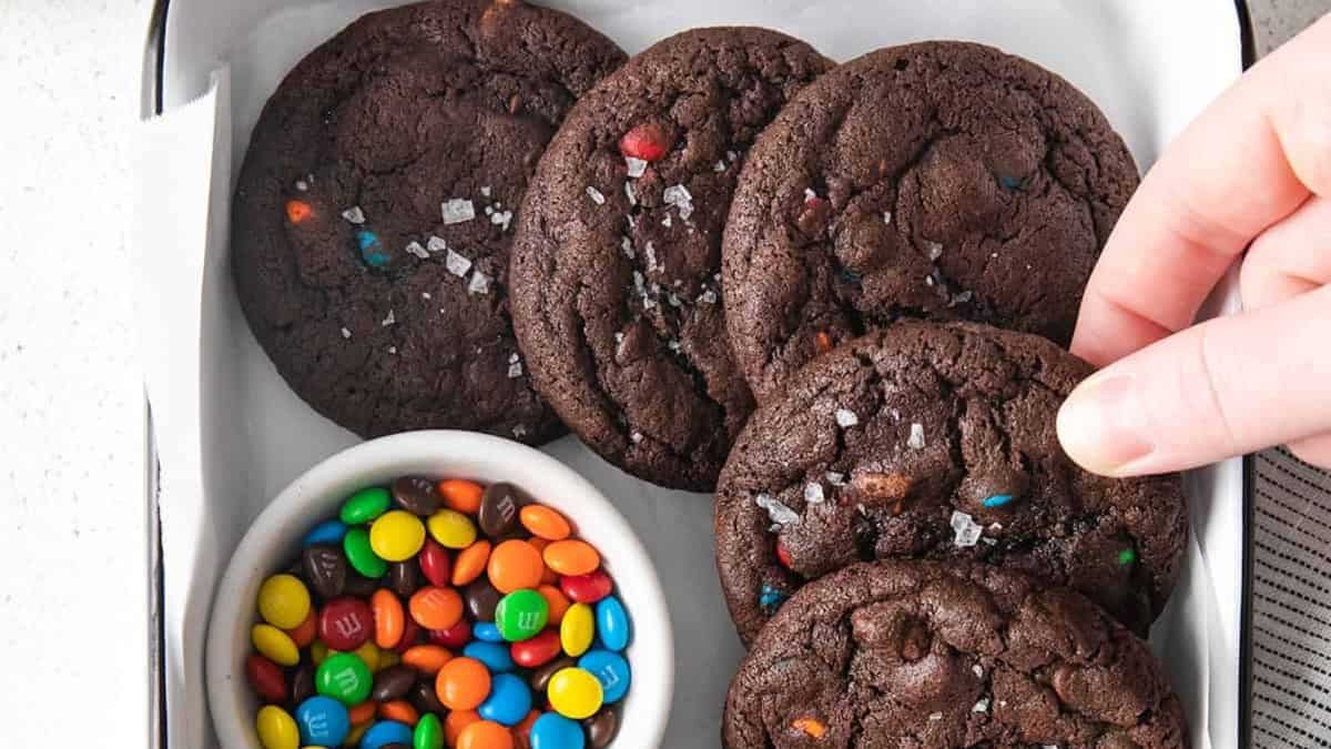 A hand reaches for a chocolate cookie with colorful candy pieces beside a small bowl of mini chocolate candies in a white tray&mdash;perfect inspiration for easy cookie recipes using pantry staples.