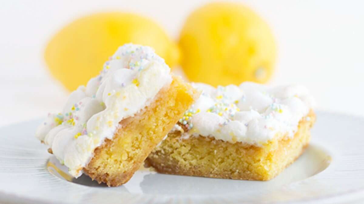 Two lemon bars with white frosting and colorful sprinkles are stacked on a white plate, with two whole lemons in the background—delightful lemon desserts perfect for spring.