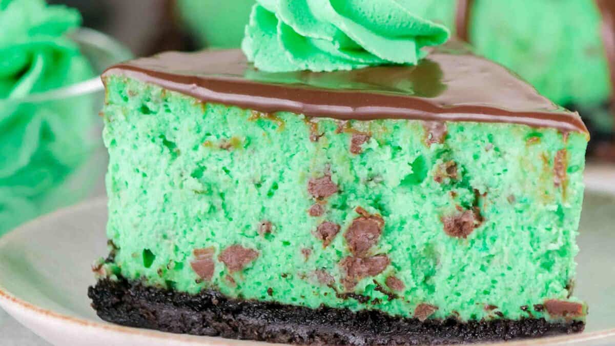 A slice of green mint chocolate chip cheesecake with a chocolate topping and cookie crust, garnished with a swirl of green whipped cream—the perfect mint chocolate dessert for St. Patrick’s Day.