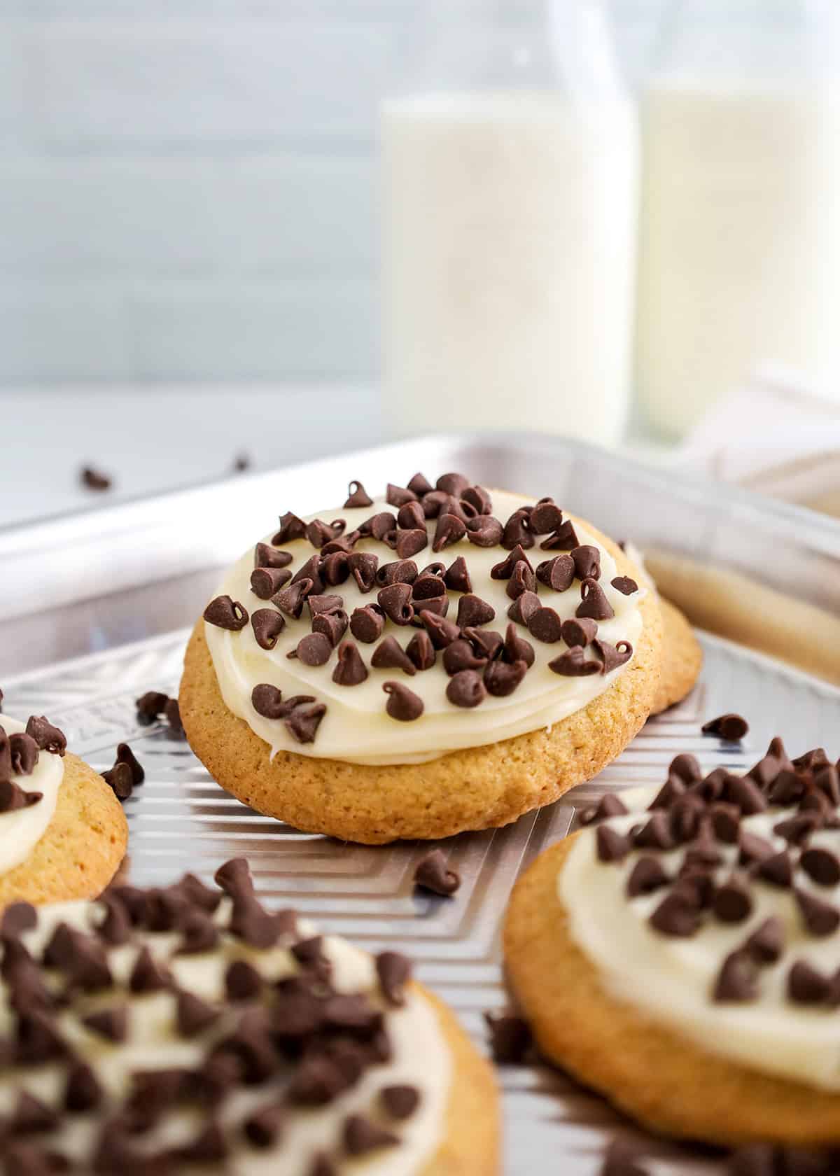 A close-up of Cannoli Cookies, frosted and topped with chocolate chips, on a baking sheet, with bottles of milk in the background.