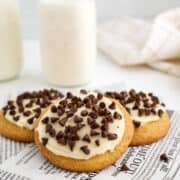 Three frosted Italian cookies topped with chocolate chips are placed on a piece of printed paper, with two bottles of milk in the background.