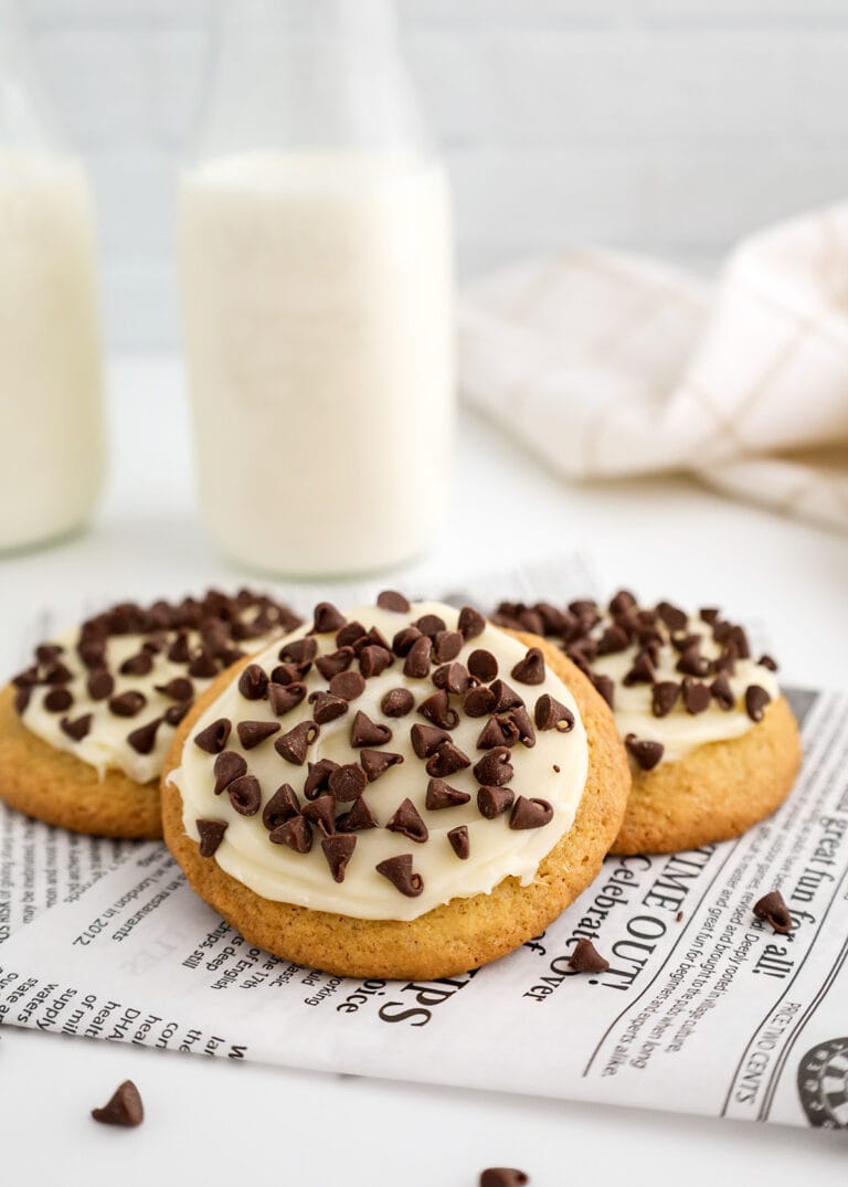 Three frosted Italian cookies topped with chocolate chips are placed on a piece of printed paper, with two bottles of milk in the background.