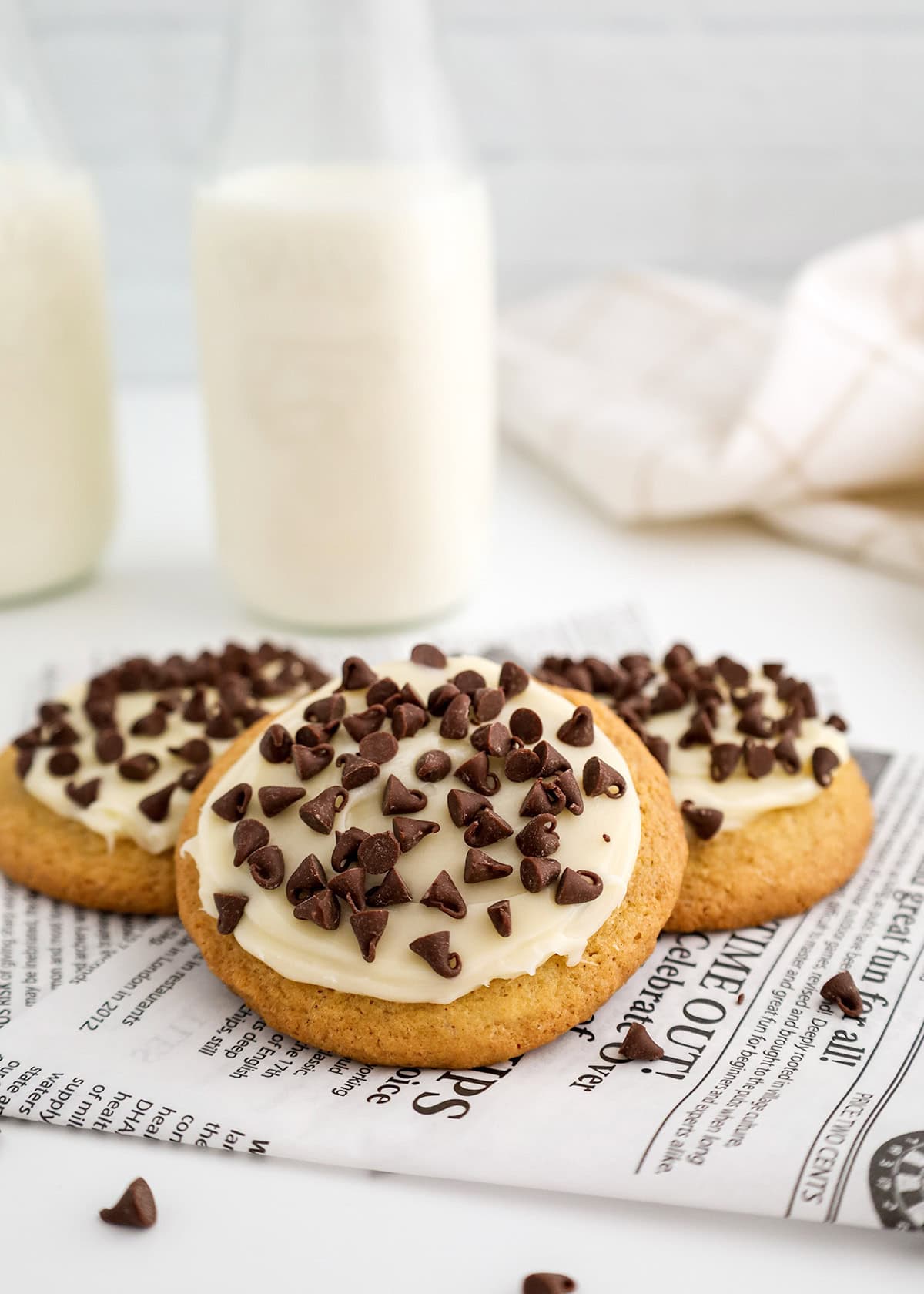 Three frosted Italian cookies topped with chocolate chips are placed on a piece of printed paper, with two bottles of milk in the background.