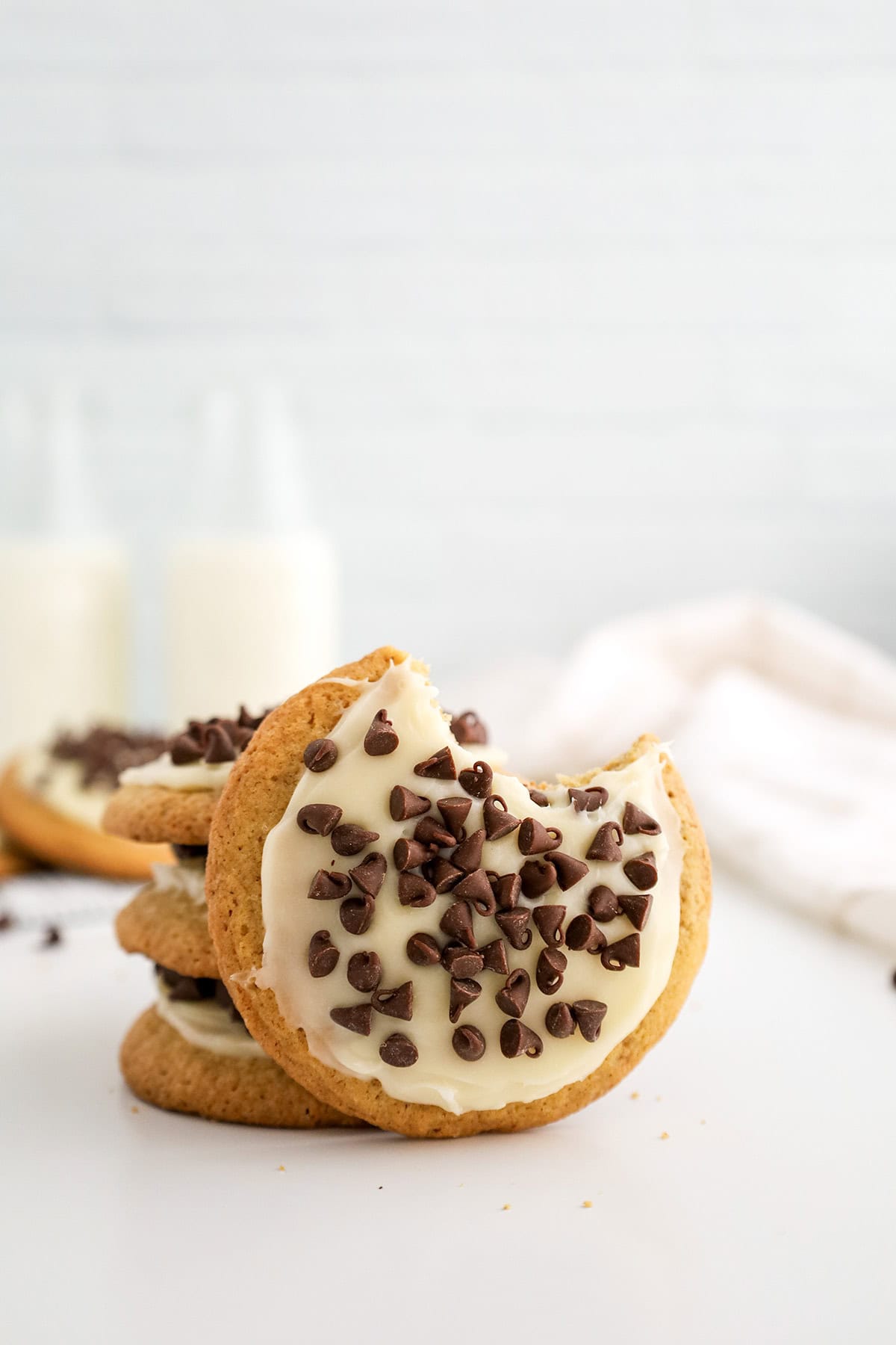 A stack of Cannoli Cookies topped with white frosting and chocolate chips, with the top cookie missing a bite; two bottles of milk are blurred in the background.