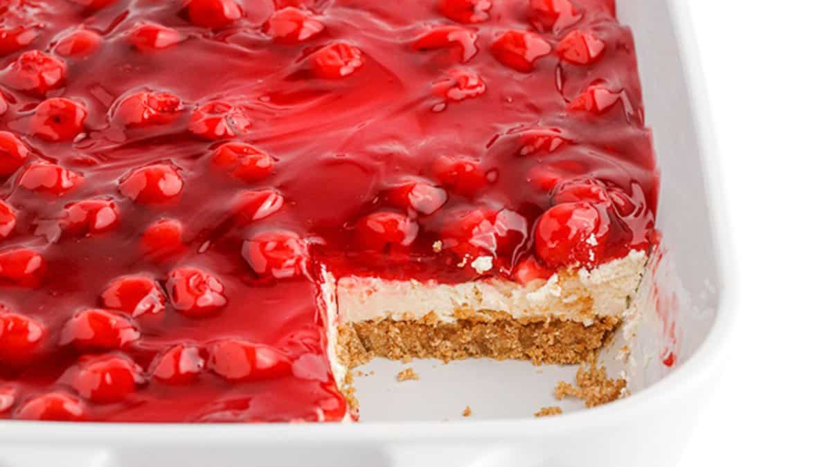 A cherry cheesecake dessert in a white dish with a slice removed, showing layers of cherry topping, creamy filling, and a graham cracker crust&mdash;one of the best no bake desserts for summer.