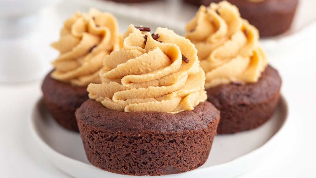 Three chocolate cupcakes with swirls of light brown frosting and chocolate sprinkles are arranged on a white plate, making them perfect mini desserts for spring parties.