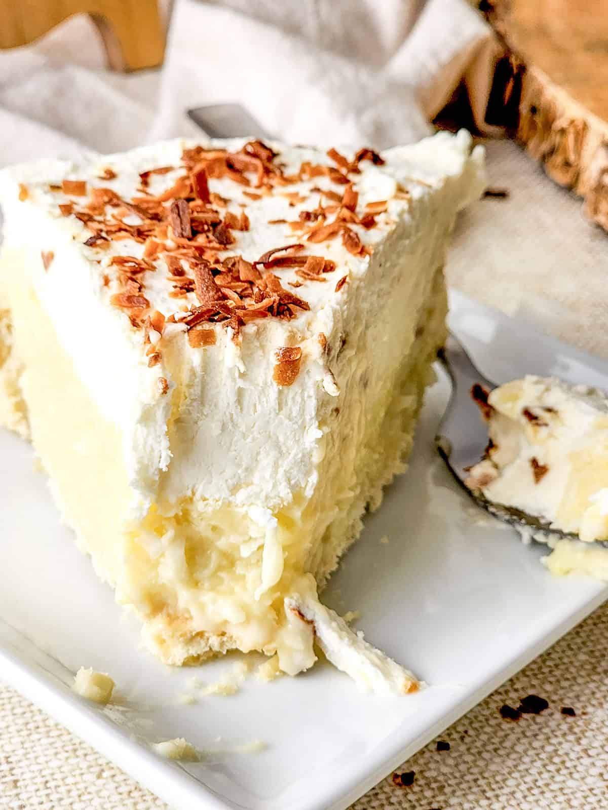 A slice of Coconut Cream Pie topped with whipped cream and toasted coconut flakes sits on a white plate, with a forkful taken out. The rich coconut filling and flaky crust make this classic dessert irresistible.