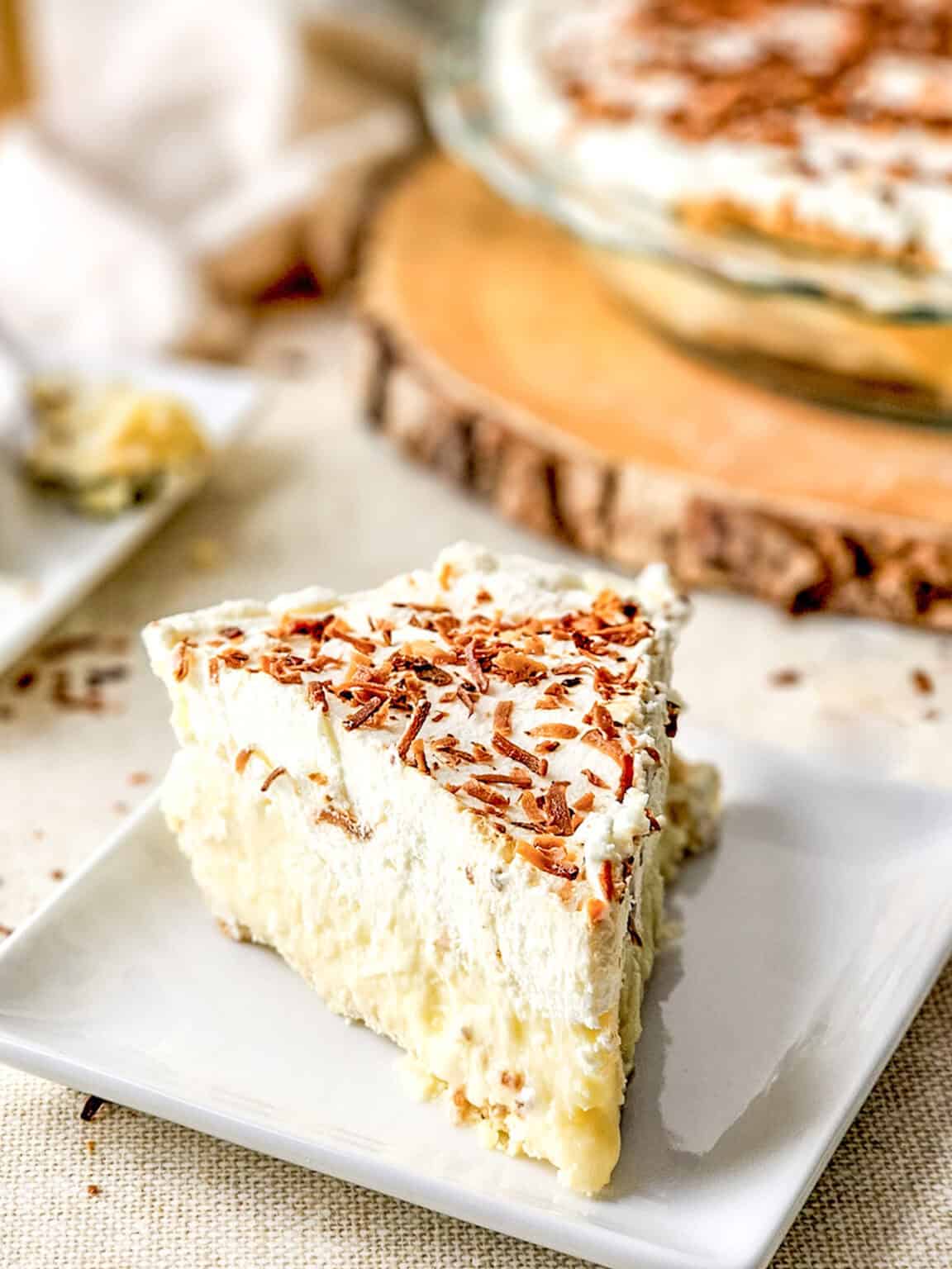 A slice of creamy Coconut Cream Pie topped with whipped cream and chocolate shavings sits on a white plate, with the rest of the pie visible in the background.