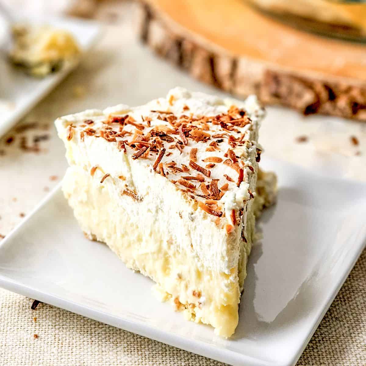 A slice of coconut cream pie topped with whipped cream and chocolate shavings sits on a white square plate.