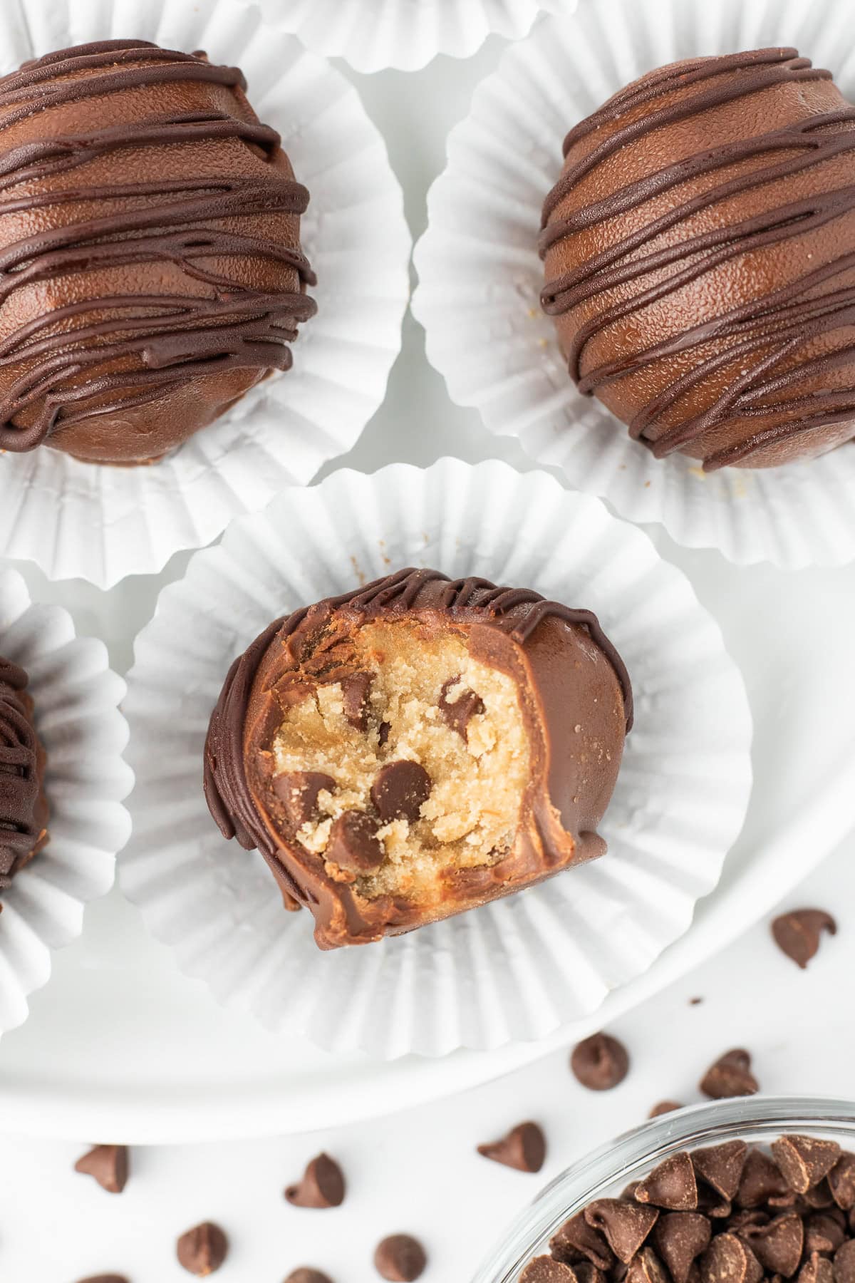 Four cookie dough truffles in paper liners, one with a bite taken out to reveal its chocolate chip cookie dough center. Chocolate chips are scattered nearby.
