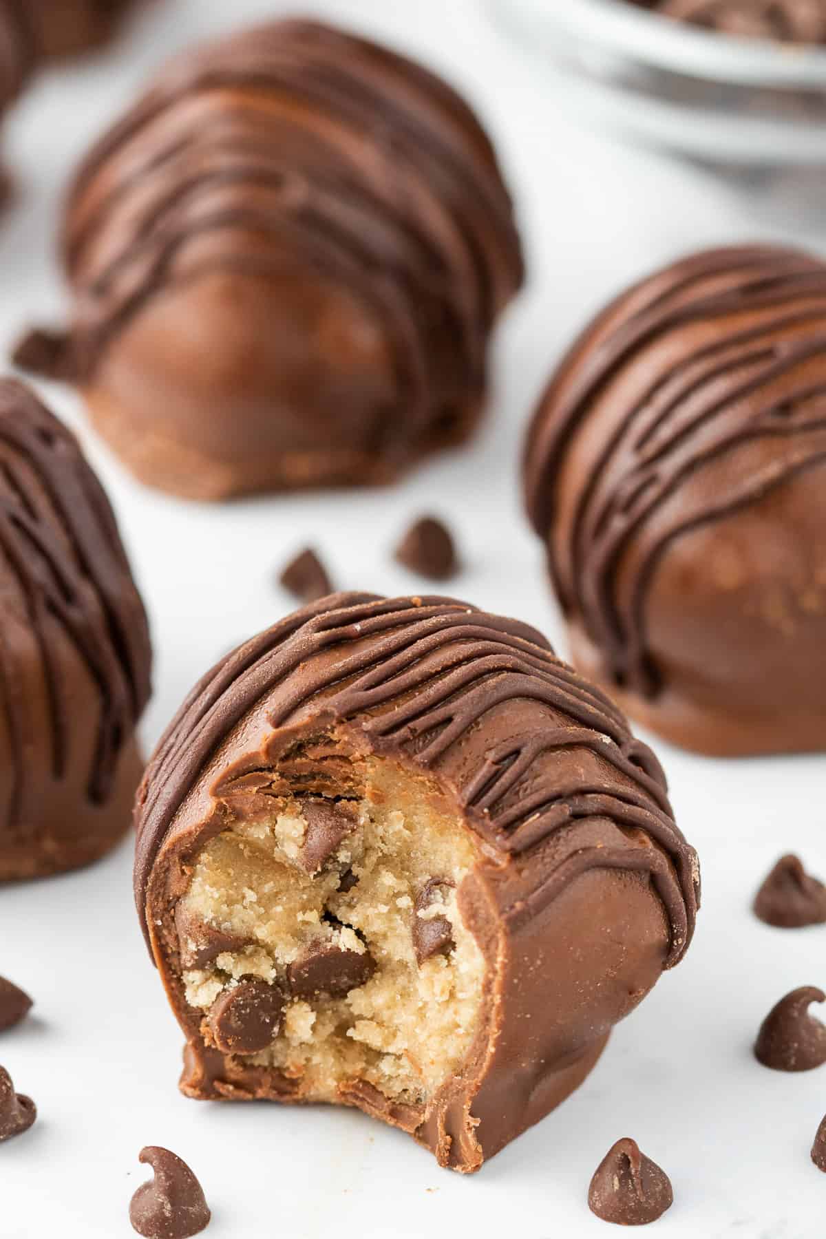 A close-up of Cookie Dough Truffles, with one truffle bitten to reveal the creamy cookie dough and chocolate chips inside. Chocolate chips are scattered nearby for an extra touch of indulgence.