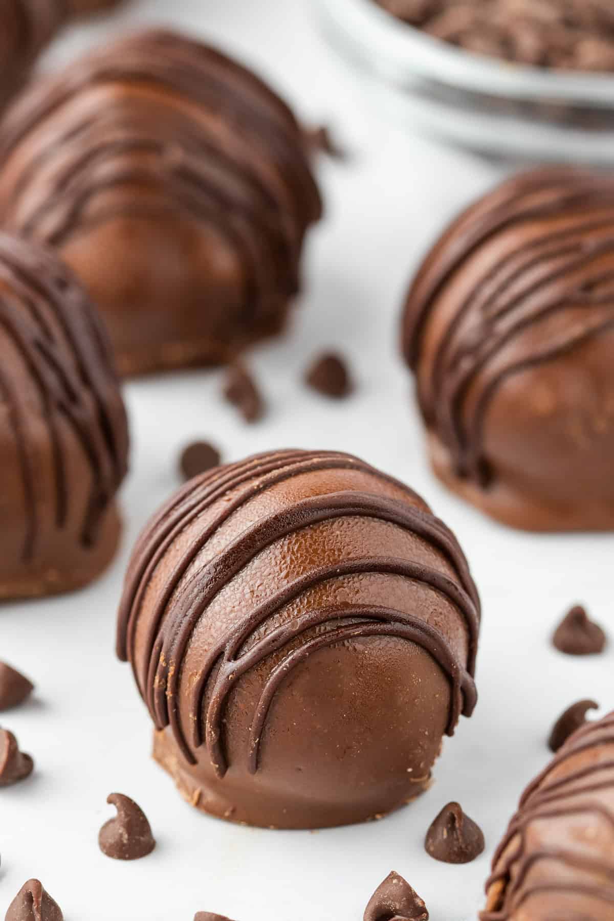 Close-up of round Cookie Dough Truffles with a drizzle of chocolate on top, surrounded by scattered chocolate chips on a white surface.