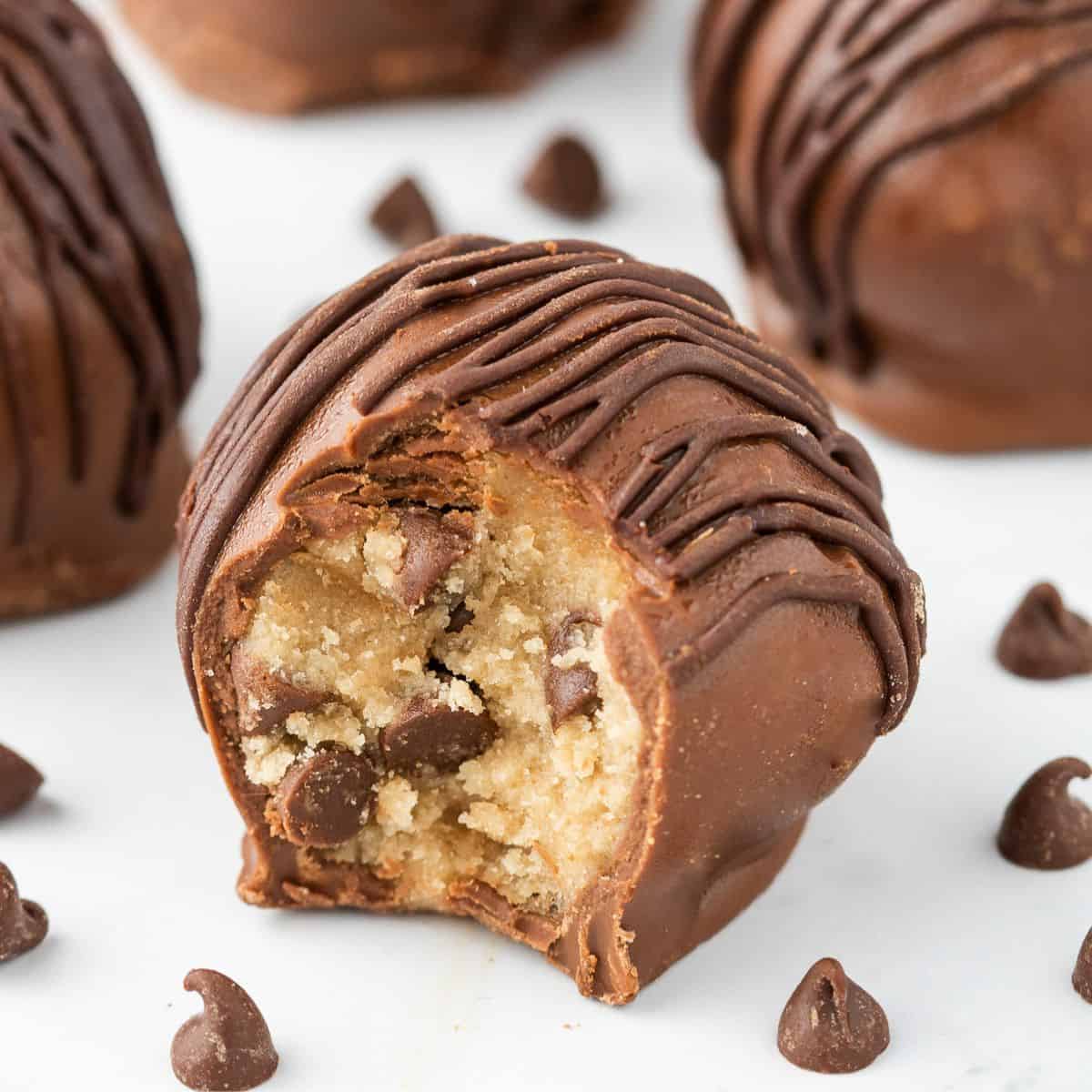 A close-up of a chocolate-coated cookie dough truffle with a bite taken out, showing the gooey cookie dough and chocolate chips inside. Cookie dough truffles and scattered chocolate chips complete the tempting scene.