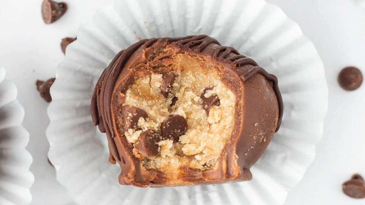 A chocolate-covered truffle with a bite taken out, revealing a cookie dough and chocolate chip filling, placed in a white paper cup—perfect for mini desserts at spring parties.