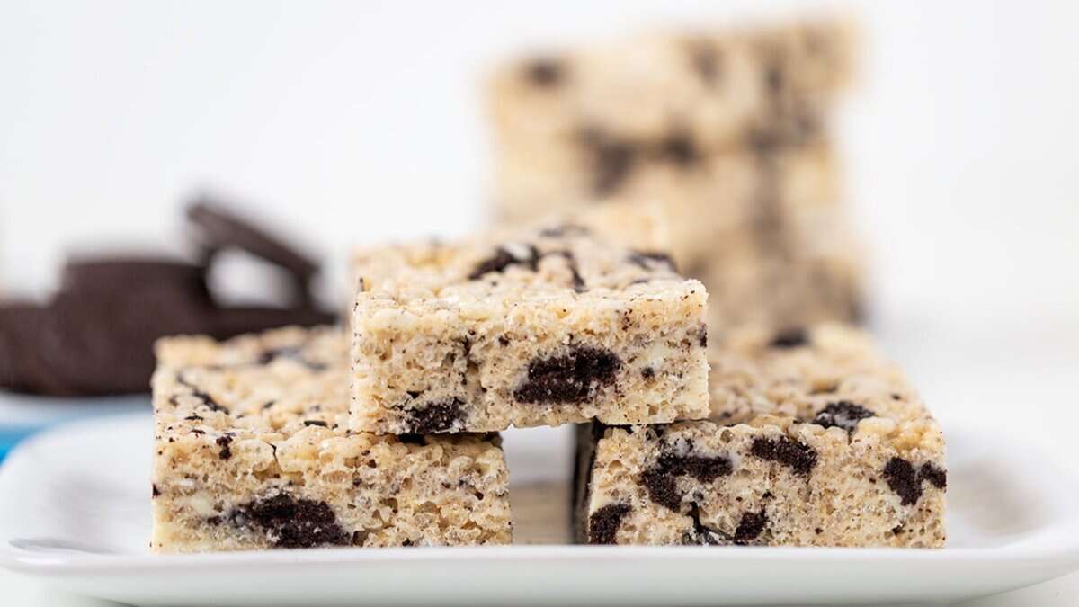 Three cereal bars with cookie pieces stacked on a white plate, with more bars and chocolate cookies blurred in the background&mdash;perfect no bake desserts for winter indulgence.