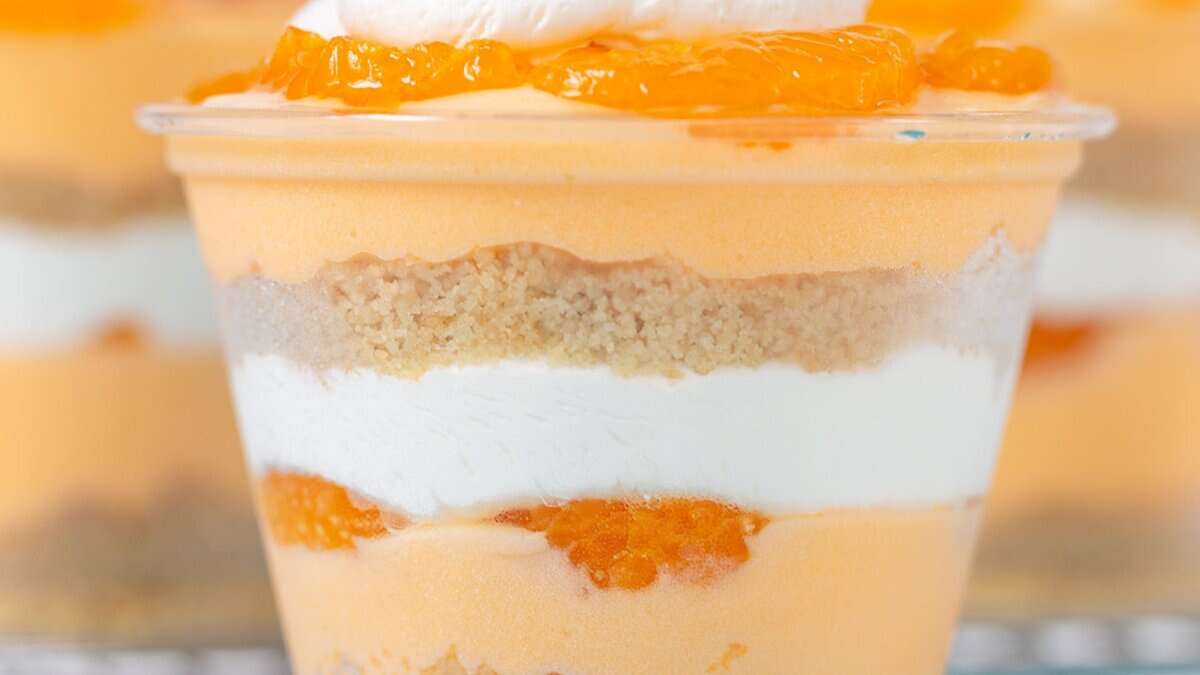 A layered mini dessert in a clear cup with orange mousse, whipped cream, crumbly crust, and mandarin orange slices—perfect for party desserts or spring parties.