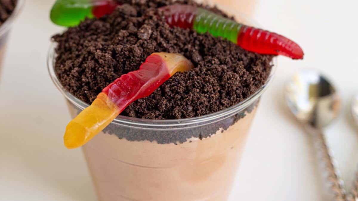 A clear plastic cup filled with chocolate pudding, topped with crushed cookies and two gummy worms, sits next to a metal spoon on a white surface&mdash;a fun and easy dessert perfect for summer desserts or no bake desserts.