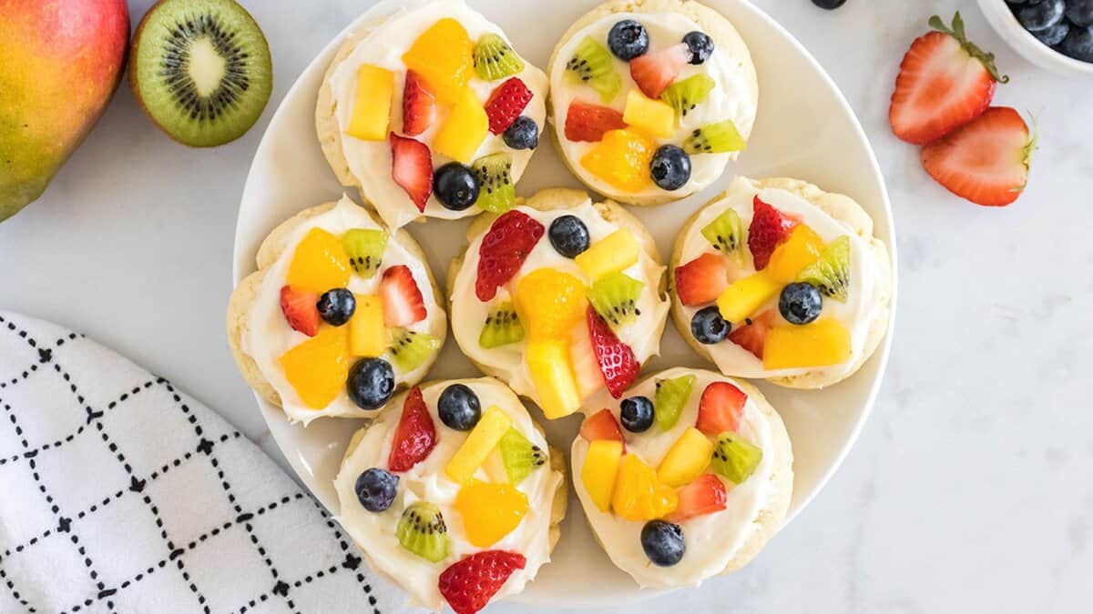 A plate of mini cookies topped with whipped cream and assorted fresh fruits like strawberries, blueberries, kiwi, and mango—perfect as mini desserts for spring parties or any festive gathering.
