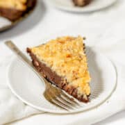 A slice of German Chocolate Pie topped with coconut and pecan frosting on a white plate with a fork, with another slice in the background.