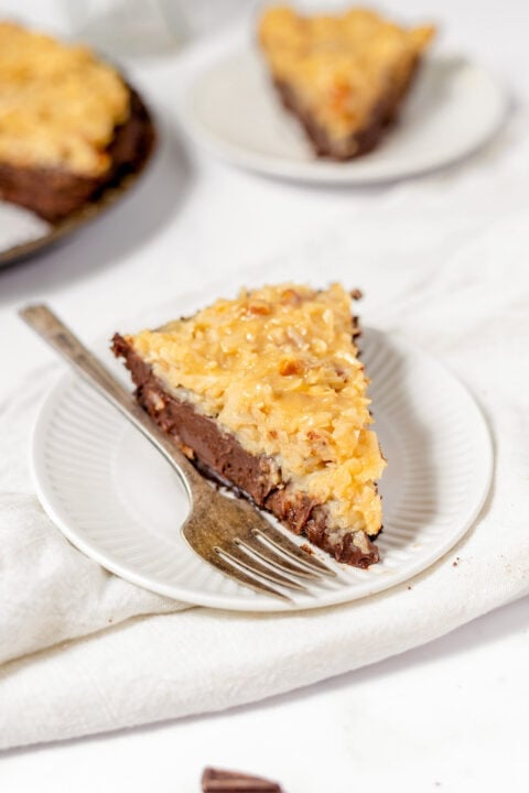 A slice of German Chocolate Pie featuring chocolate and coconut filling sits with a fork on a white plate, with another tempting slice in the background.