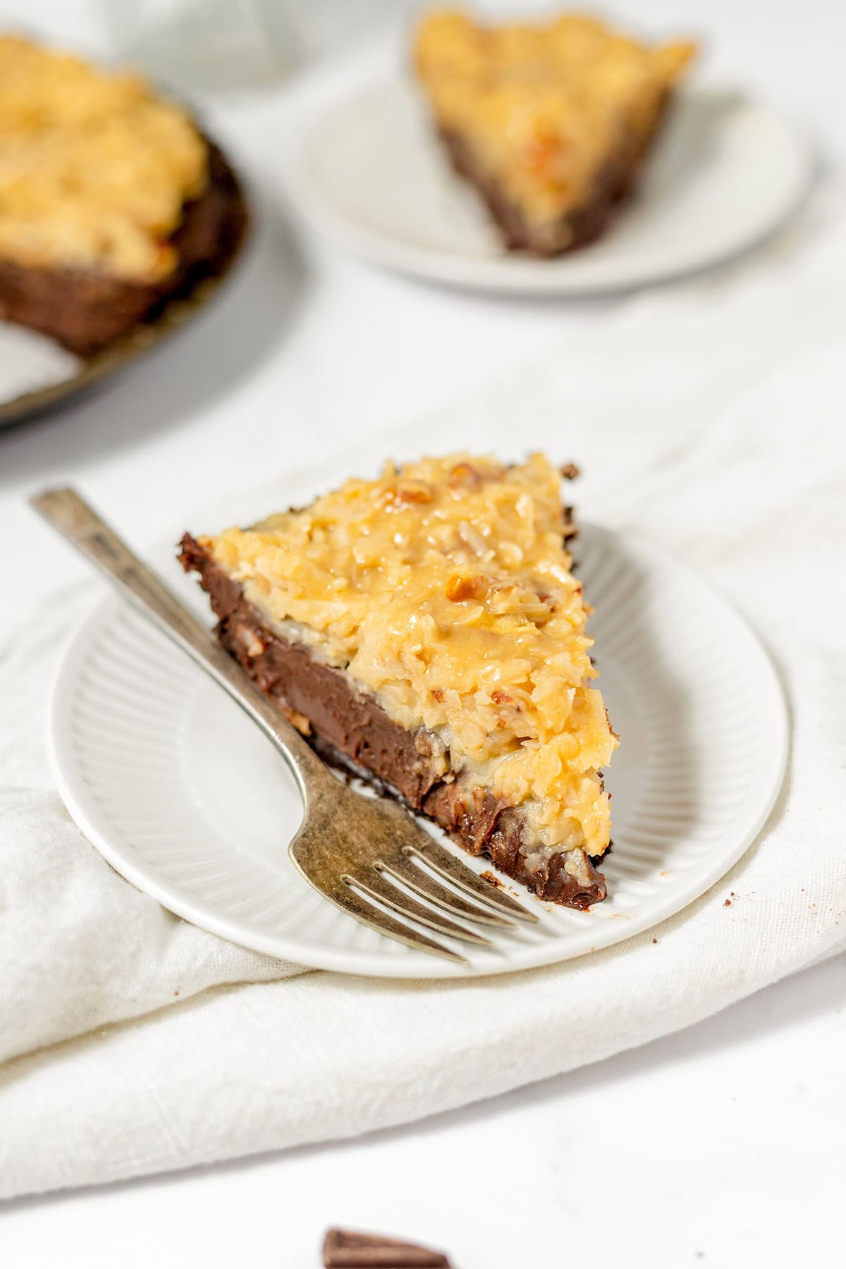 A slice of German Chocolate Pie topped with coconut and pecan frosting on a white plate with a fork, with another slice in the background.