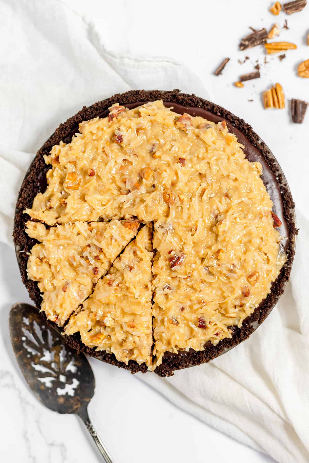 A decadent German Chocolate Pie with a coconut and pecan topping, partially sliced on a marble surface, pie server ready and scattered pecan pieces nearby.
