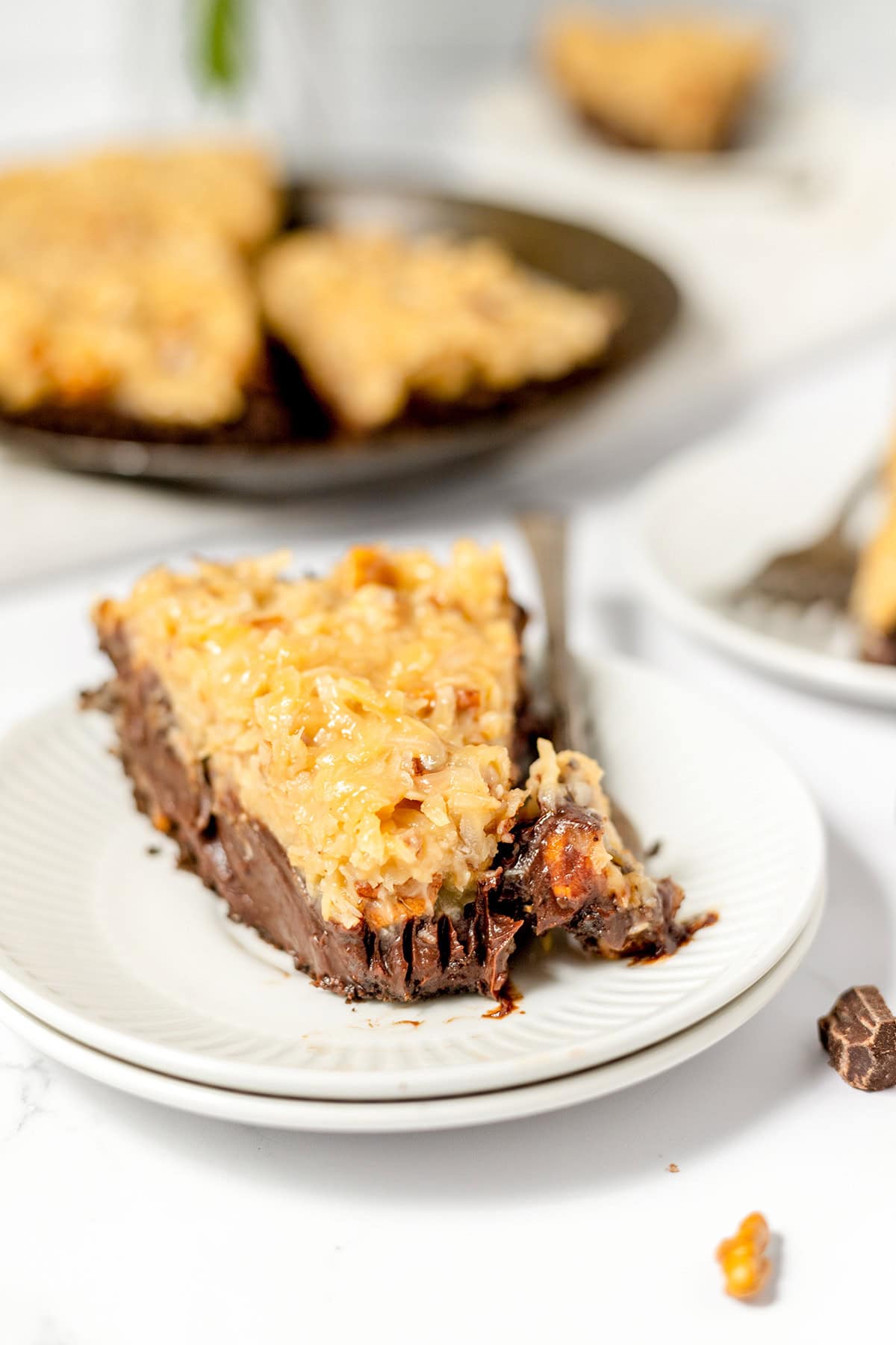 A slice of German Chocolate Pie topped with a coconut and pecan mixture sits on a white plate, with a fork and more pie slices in the background.