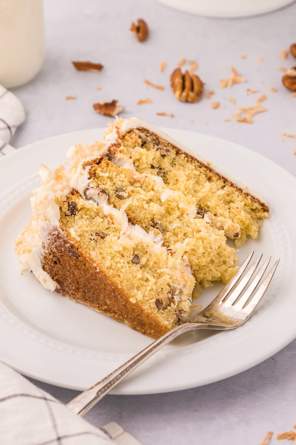 A slice of three-layer Italian Cream Cake with white frosting on a white plate, garnished with shredded coconut and pecans, with a fork beside it.