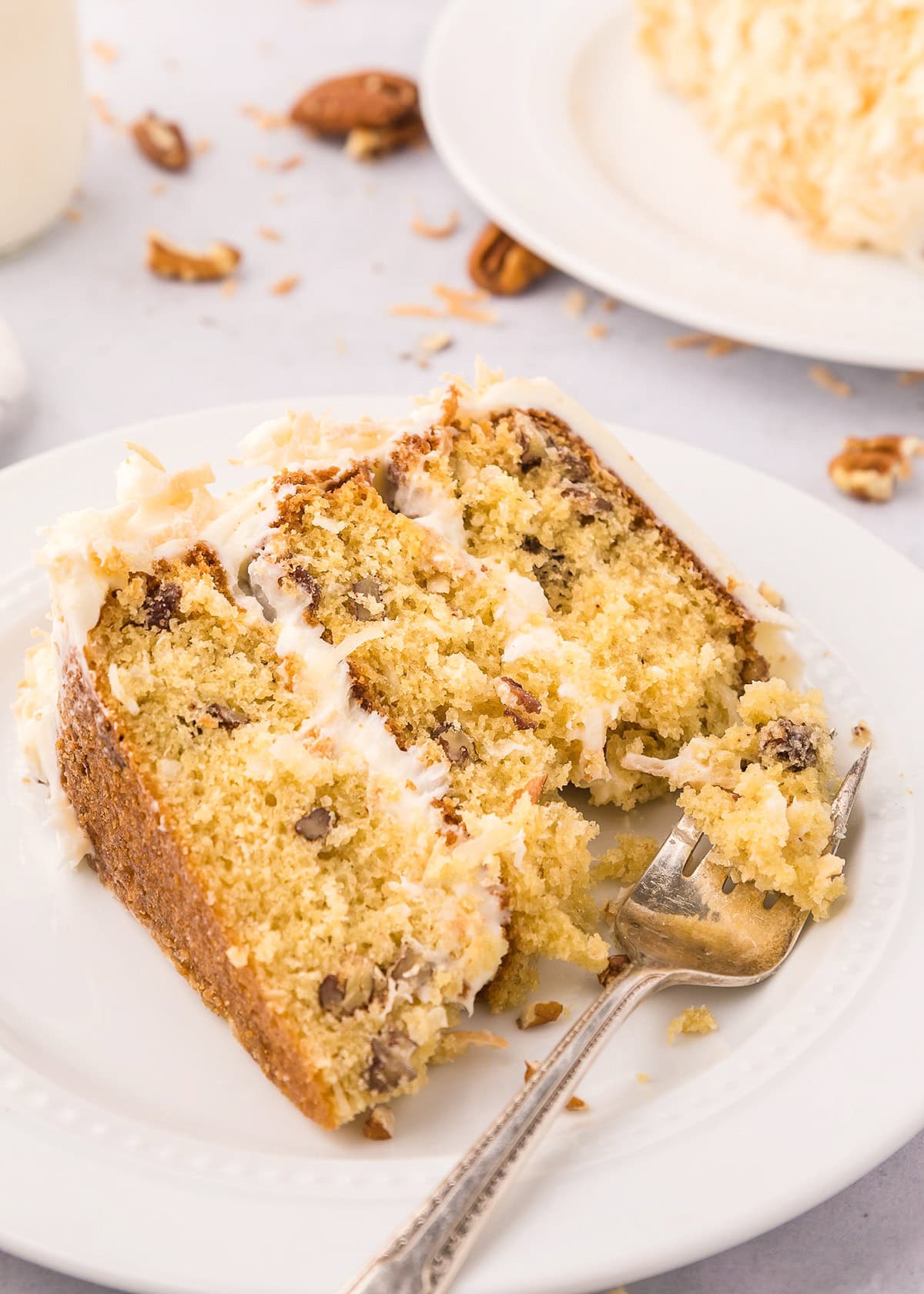 A slice of Italian Cream Cake with frosted layers and pecans is partially eaten on a white plate with a fork, cake crumbs and nuts scattered around.