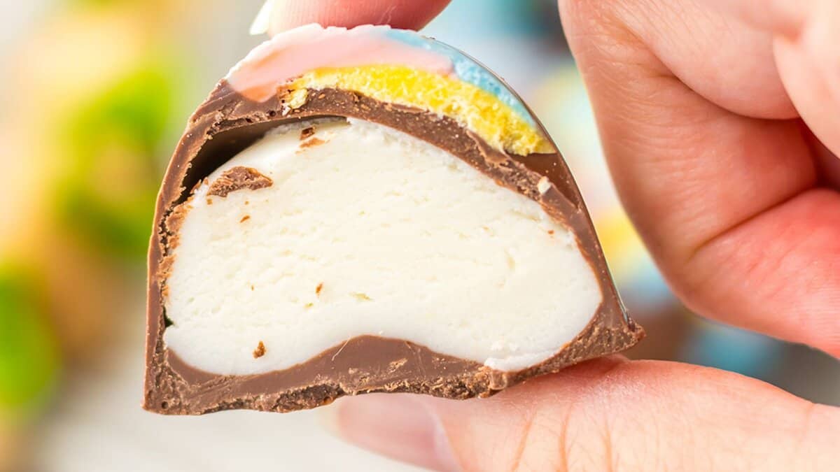 A hand holding a cross-section of a chocolate-coated confection with a creamy white filling and colorful top layer&mdash;perfect for holiday desserts or as a delightful addition to your favorite Easter recipes.