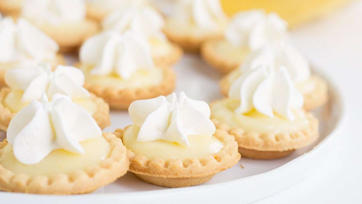 Mini desserts perfect for spring parties—these tartlets feature a yellow custard filling and a swirl of whipped cream, beautifully arranged on a white plate.