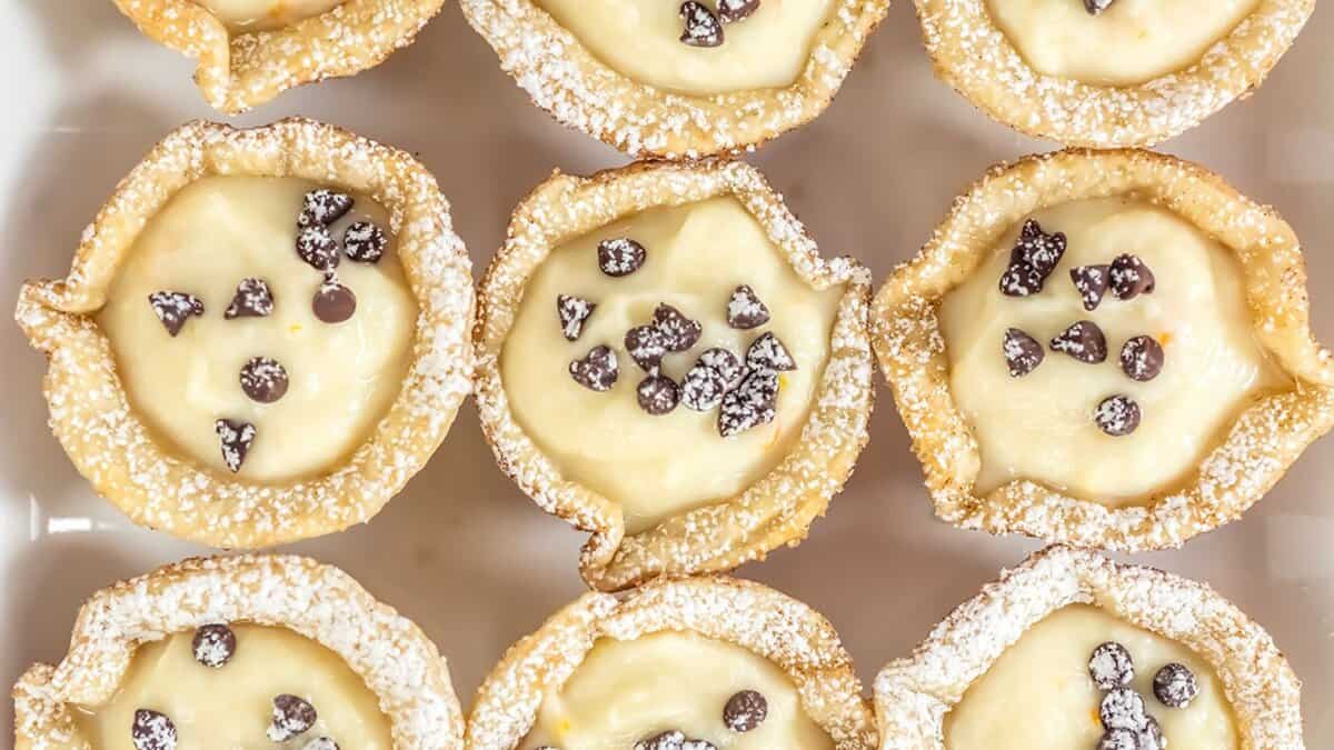 Nine mini cannoli cups filled with cream and topped with mini chocolate chips, arranged in a grid and dusted with powdered sugar—perfect for spring parties or as delightful mini desserts at any gathering.