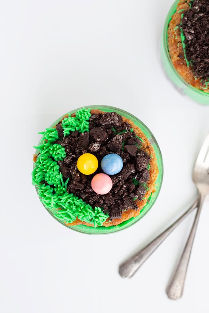 A Mini Easter Trifle dessert cup topped with green icing, crumbled cookies, and three colorful candy eggs, with two spoons beside it on a white surface&mdash;a festive addition to your collection of Easter desserts.